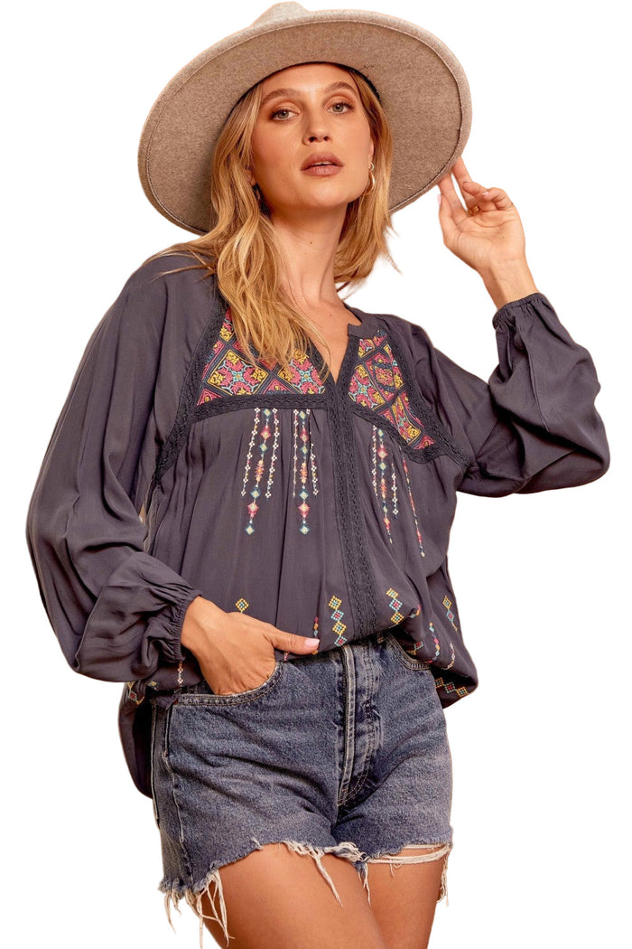 A woman wearing a navy blue embroidered lace blouse with a split V-neck, long balloon sleeves, and lace detailing. She is also wearing a wide-brimmed hat and cut-off denim shorts.