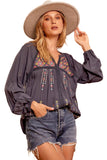 A woman wearing a navy blue embroidered lace blouse with a split V-neck, long balloon sleeves, and lace detailing. She is also wearing a wide-brimmed hat and cut-off denim shorts.