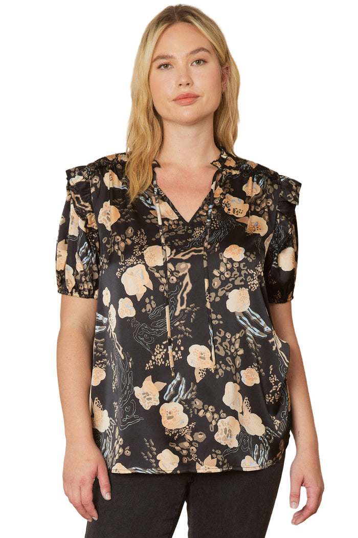 A woman wearing a black floral and ruffle blouse with a V-neck, short puffed sleeves, and ruffle detailing.