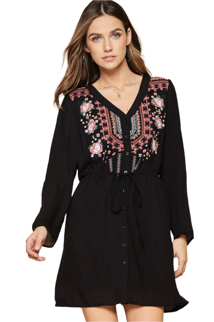 A woman wearing a black embroidered tunic dress with a V-neckline, button down front, and roll tab sleeves.